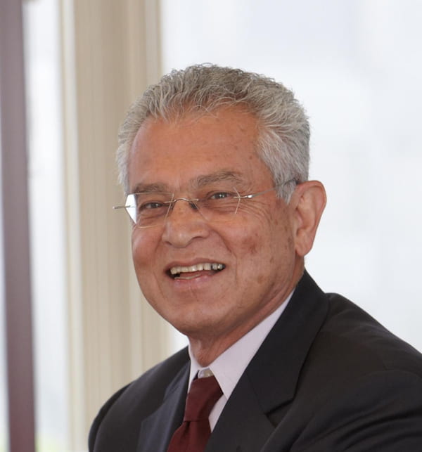 This image shows Dr. Bahman Sheikh, a smiling middle-aged man with grey, short, slightly wavy hair. He is wearing rectangular, thin-framed glasses and a dark suit with a light-colored shirt. The background is bright and appears to be an indoor setting with natural light. The man exudes a professional and friendly demeanor.